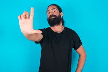 young bearded man wearing black T-shirt over blue studio background making fun of people with fingers on forehead doing loser gesture mocking and insulting.