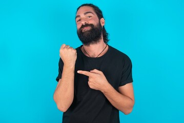 young bearded man wearing black T-shirt over blue studio background In hurry pointing to wrist watch, impatience, looking at the camera with relaxed expression