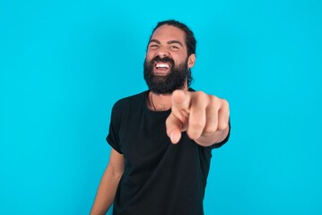Naklejka premium young bearded man wearing black T-shirt over blue studio background pointing displeased and frustrated to the camera, angry and furious ready to fight with you.