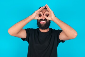 young bearded man wearing black T-shirt over blue studio background doing ok gesture like binoculars sticking tongue out, eyes looking through fingers. Crazy expression.