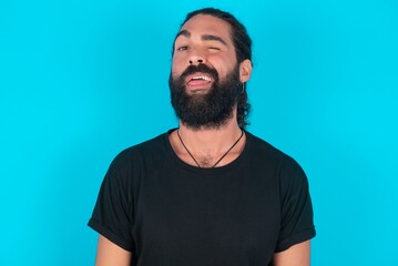 young bearded man wearing black T-shirt over blue studio background sticking tongue out happy with funny expression. Emotion concept.