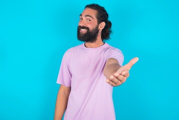 Fototapeta premium young bearded man wearing violet T-shirt over blue studio background smiling friendly offering handshake as greeting and welcoming. Successful business.