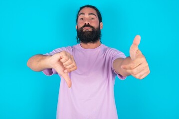 young bearded man wearing violet T-shirt over blue studio background showing thumbs up and thumbs down, difficult choose concept - obrazy, fototapety, plakaty