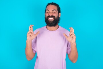 young bearded man wearing violet T-shirt over blue studio background has big hope, crosses fingers,...