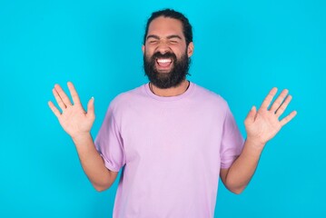 Emotive young bearded man wearing violet T-shirt over blue studio background laughs loudly, hears...