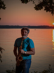 An adult man plays the saxophone at sunset by the river in the evening
