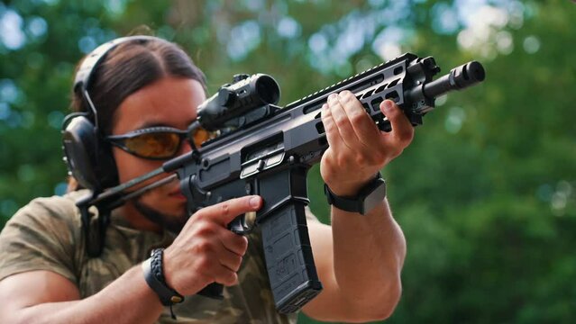 White muscular man in camouflage t-shirt and safety gear firing submachine gun on training ground. Outdoor horizontal shot. High quality 4k footage