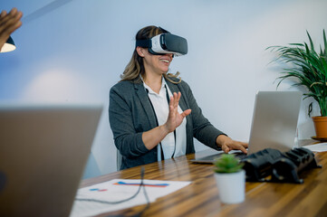 Happy female employee in VR goggles typing on laptop in office