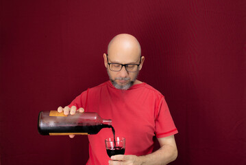 Young handsome man with beard, bald, red shirt wearing glasses holding in his hands a bottle of grape juice and a glass glass against red background.