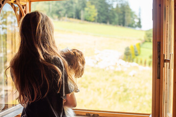 Rearview of long-haired brunette woman with dog in hands looking out the window, contemplating landscape rural nature