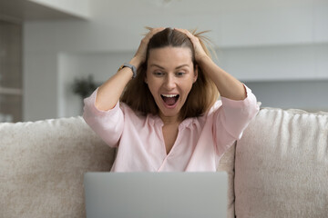 Excited cheerful pretty 30s laptop user woman staring at display, shouting for joy, gasping, laughing, holding head in amazement, surprise, getting good awesome news, sitting on home couch