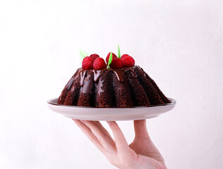 Hand holding chocolate cake with fluted sides topped with glazing and raspberry on ceramic plate. Serving appetizing dessert