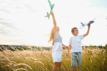 Running boy and girl holding two green and blue airplanes toy in the field during summer sunny day