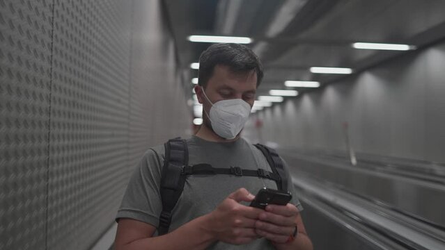 A young guy wearing a protective mask on public transportation descends the subway escalator and uses his smartphone. Focused man with a covid mask on an elevator in the metro, surfing the Internet