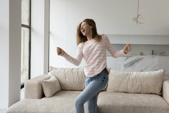 Happy excited pretty young woman having fun at home, enjoying energetic activity, home party, dancing to music at couch, jumping, singing songs, laughing, celebrating real estate buying