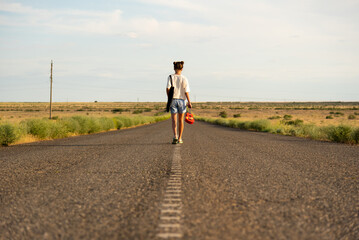 Obraz premium A girl walks along an empty road in the steppe. She has a ukulele in her hands and a backpack on her shoulders. It's summer and warm outside. Open road.