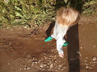 Little toddler baby boy in rubber boots playing in the puddle with mud in summer. Top view of the walking child.