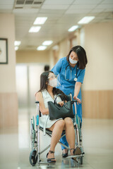 Smiling young female doctor in scrub and covid-19 face mask in conversation with patient sitting in wheelchair holding purse during discharge from hospital