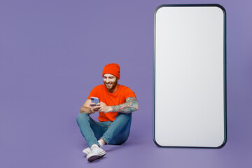 Full body smilinng fun young man in red hat tshirt use smartphone sit near big huge blank screen mobile cell phone workspace mockup isolated on plain pastel purple background People lifestyle concept