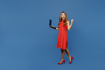 Full body side view young IT woman 30s wear red dress hold use work on laptop pc computer do winner gesture isolated on plain dark royal navy blue background studio portrait. People lifestyle concept.