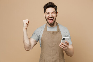 Young happy man barista barman employee in brown apron work in coffee shop hold use mobile cell...