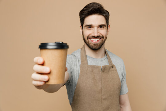 Young Cheerful Man Barista Barman Employee Wear Brown Apron Work In Shop Hold Give Craft Paper Brown Cup Coffee To Go Isolated On Plain Pastel Light Beige Background. Small Business Startup Concept.