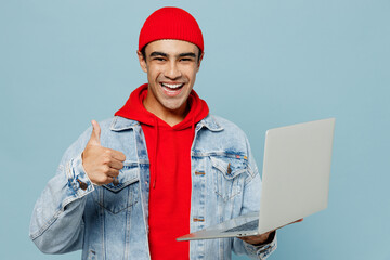 Young fun middle eastern IT man 20s he wear denim jacket red hat hold use work on laptop pc computer show thumb up isolated on plain pastel light blue cyan background studio. People lifestyle concept.