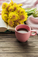 Morning coffee. A bouquet of yellow dandelions on an open book and a cup of coffee on a wooden table. Cozy morning. Copy space.