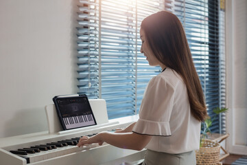 Side view of focused young Asian female practicing playing on white electric piano at home studio...