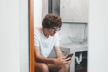 Fototapeta na wymiar Young Caucasian man gamer sitting on toilet bowl in restroom playing game online on mobile smartphone gadget. Addiction dependency habit social network media technology. Man using phone.