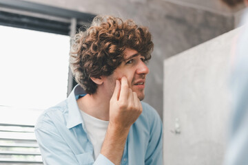 Portrait of young Caucasian man using his hand squeezed painful inflammatory acne on the cheek with. Skincare beauty concept. Handsome man looking at himself in the bathroom mirror Squeezing pimple.