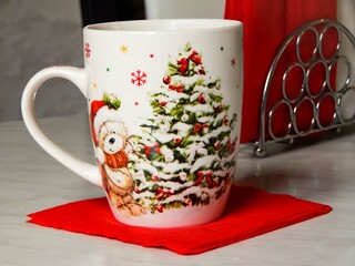 A Christmas mug with a picture of a Christmas tree stands on a red napkin, a napkin holder with white and red napkins in the background