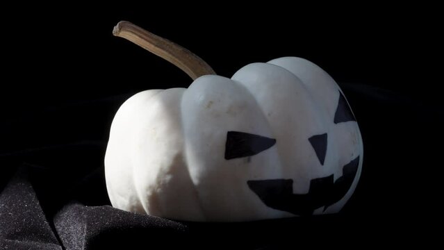 A white pumpkin decorated for Halloween rotates on a black background. Ray of the sun