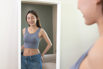 Asian woman wearing her old pant before loss weight looking reflection in the mirror and feeling joyful with her body after diet success