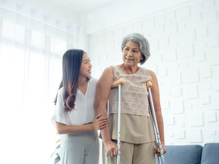 Fototapeta na wymiar Elderly woman trying to walk on crutches standing held and supported in arms by young Asian female carefully in recovery room, helping old women, health care, senior therapy patient at home concept.