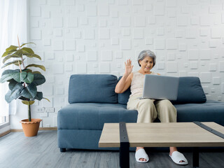 Happy Asian senior casual woman sitting on the sofa, waving greeting at laptop computer screen in white room. Elderly female meet with family by video call at home. Older people with technology.