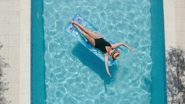 4K footage over swimming pool with relaxing woman in black swimsuit, straw hat drifting on inflatable mattress on the blue ripples water surface. Holidays, recreation concept.