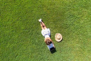 Woman in a white dress, hat with laptop, lying on the green grass at summer day. Top view, drone,...