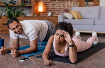 Fototapeta premium Smiling young black man supports tired sad wife in sportswear do plank exercises on mat