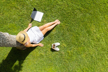 Woman in a white dress, hat with laptop, sitting on the green grass under tree at summer day. Top...