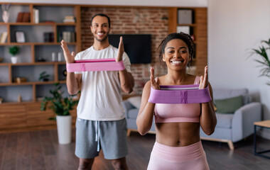 Smiling young african american husband and wife in sportswear doing arm exercises with rubber band