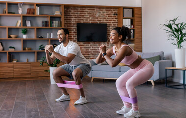 Happy young african american man and lady in sportswear do squat and leg exercises with rubber band