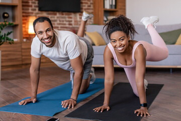 Fototapeta na wymiar Glad young african american male and female in sportswear doing leg exercises on mat in living room