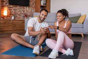 Cheerful young african american husband and wife in sportswear watch video on smartphone on mat