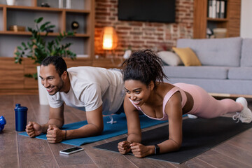 Smiling young black female and male in sportswear doing plank exercises together on mat