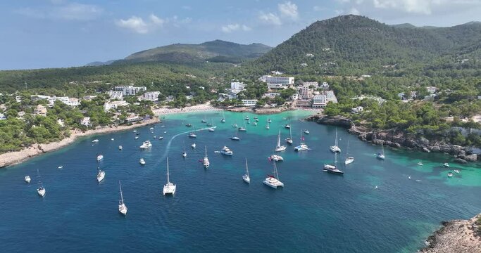 Paradise beach surrounded by trees. Yachts and boats anchored in front of the shore where people enjoy the sun.