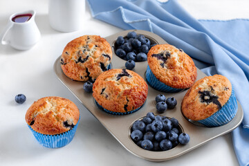 Freshly baked homemade blueberry muffins in a muffin tin with heap of fresh blueberries and sweet berry syrup on a white background.