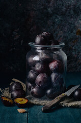 A glass jar with dark red plums stands on a blue wooden table, next to it are several plums, one cut and a small old knife