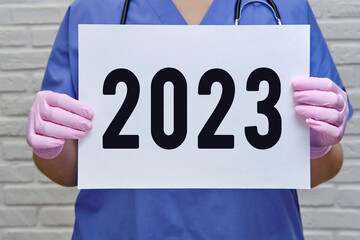 A female doctor holds an empty blank sheet, text 2023 year. Hands of a nurse in protective medical...