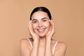 Beautiful young woman with cream on skin under eye against beige background